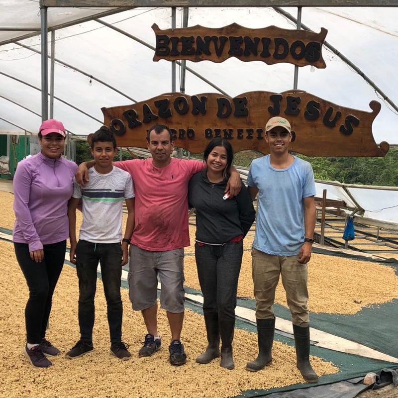 Five people standing together inside a greenhouse or barn structure under a large wooden sign that reads 'BIENVENIDOS' and 'ORACION DE JESUS'. The ground is covered with yellow grains or seeds, and the background shows the white translucent roof of the structure.