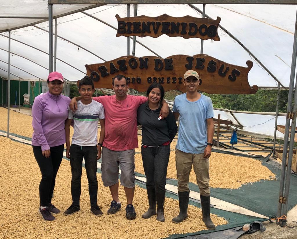 Five people standing together inside a greenhouse or barn structure under a large wooden sign that reads 'BIENVENIDOS' and 'ORACION DE JESUS'. The ground is covered with yellow grains or seeds, and the background shows the white translucent roof of the structure.
