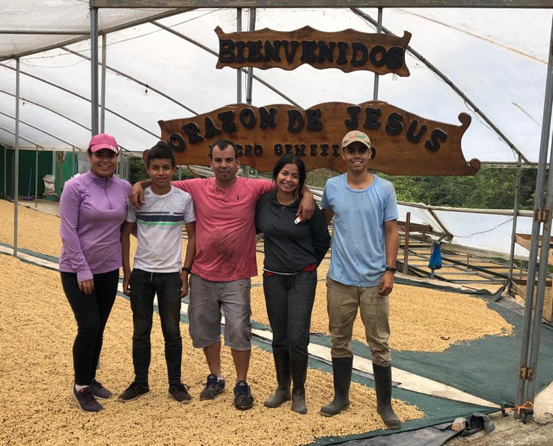 Five people standing together inside a greenhouse or barn structure under a large wooden sign that reads 'BIENVENIDOS' and 'ORACION DE JESUS'. The ground is covered with yellow grains or seeds, and the background shows the white translucent roof of the structure.