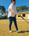Woman raking a field of coffee cherries to dry, with bright yellow building in the background and vibrant blue sky